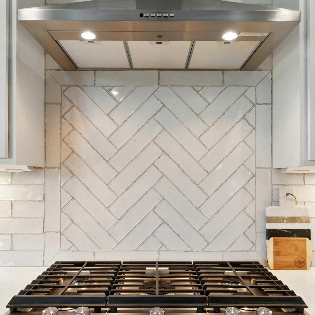 Clean white herringbone backsplash with a subtle sheen, installed as a full-height focal point behind a stainless range—great for modern or creative kitchen styles.
